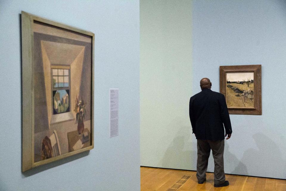 In this Monday, Feb. 6, 2017 photo, Niles Spencer's 1927 painting "The Dormer Window," left, and Andrew Wyeth's 1946 painting "The Stone Fence" are displayed as a security guard walks through the exhibit "Cross Country: The Power of Place in American Art, 1915-1950," at the High Museum of Art in Atlanta. The new exhibition at takes a look at how American artists during the modernist period traveled outside cities to find inspiration in the rural landscape. (AP Photo/David Goldman)