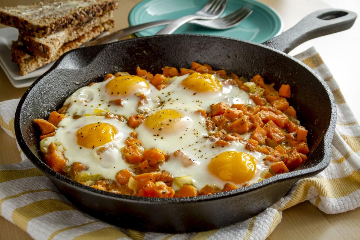 Fried eggs and sweet potato hash in cast iron skillet sitting on yellow striped kitchen towel with whole grain toast and stacked plates