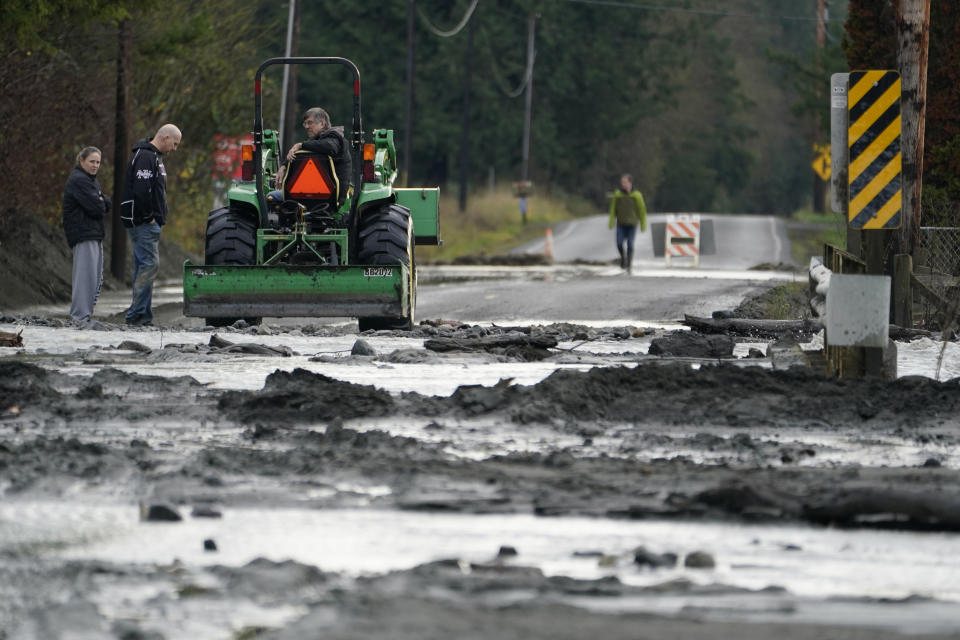 A tractor operator talks with people standing near a mud- and debris-covered road near Everson, Wash., Monday, Nov. 29, 2021. Localized flooding was expected Monday in Washington state from another in a series of rainstorms, but conditions do not appear to be as severe as when extreme weather hit the region earlier in November. (AP Photo/Elaine Thompson)
