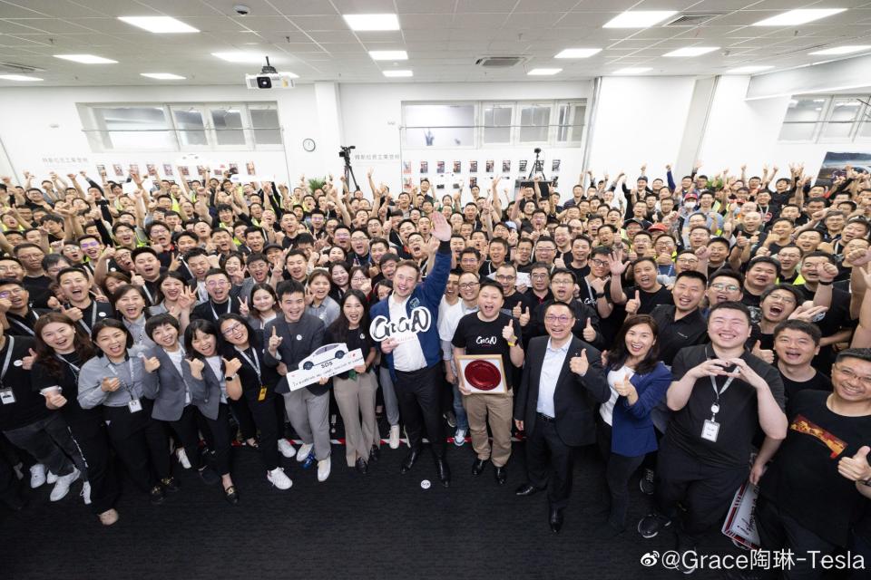 Elon Musk holding a sign and waving while standing with a large group of workers at Tesla's Shanghai gigafactory.