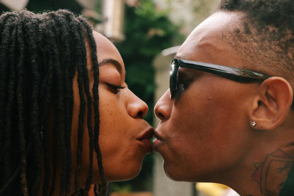 Two people stand with their lips very close together, one blowing smoke into the other's mouth.