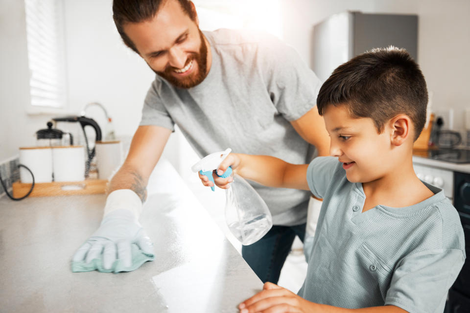 Helping, learning and teaching young son how to clean family home having fun and bonding together. Smiling and loving father cleaning, washing and wiping kitchen counter with his little boy indoors