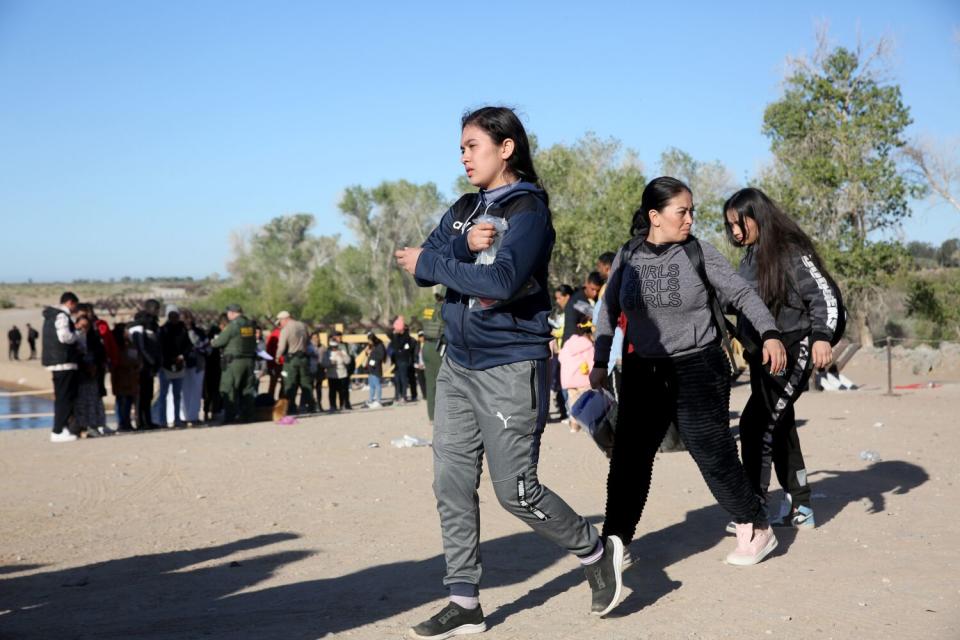 A teenage girl walking in the sun with others as a larger group waits in the background near a grove of trees