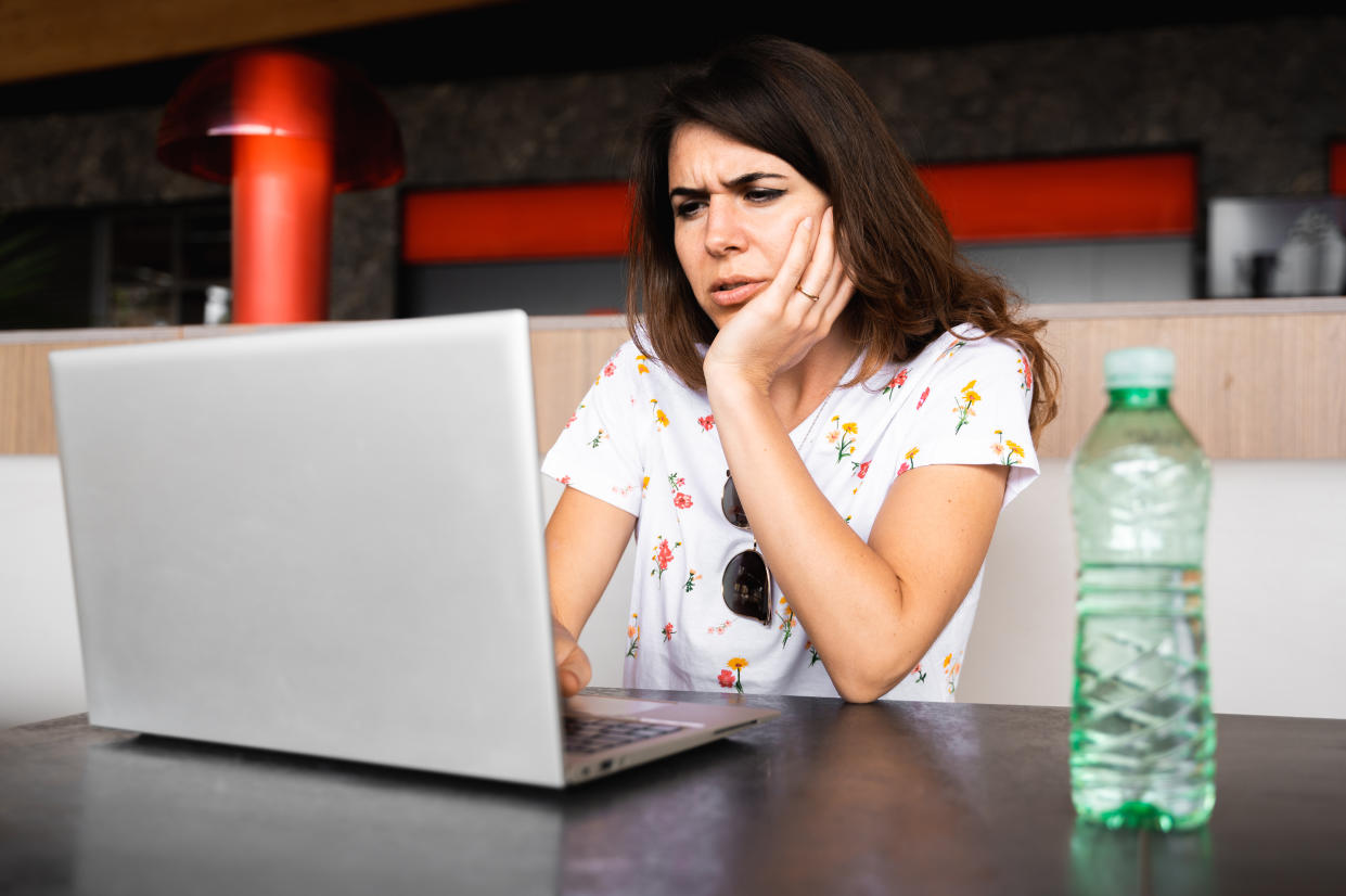 Casual young woman using computer notebook in a public place. Female with serious confused expression supports cheek by hand.