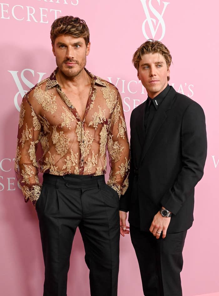 Two men posing, one in a sheer lace shirt and the other in a black suit, at an event