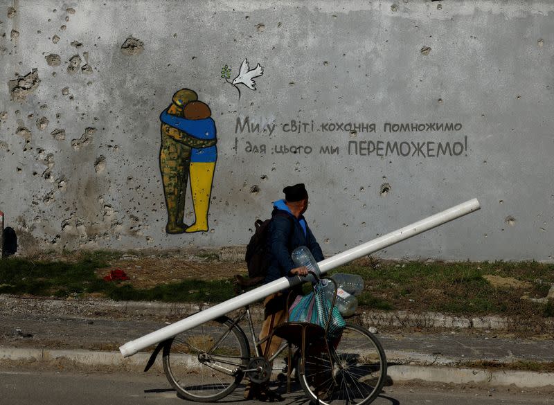 Un hombre pasa en bicicleta junto a un grafiti en la pared de un edificio cubierto con restos de balas y metralla en Kupiansk