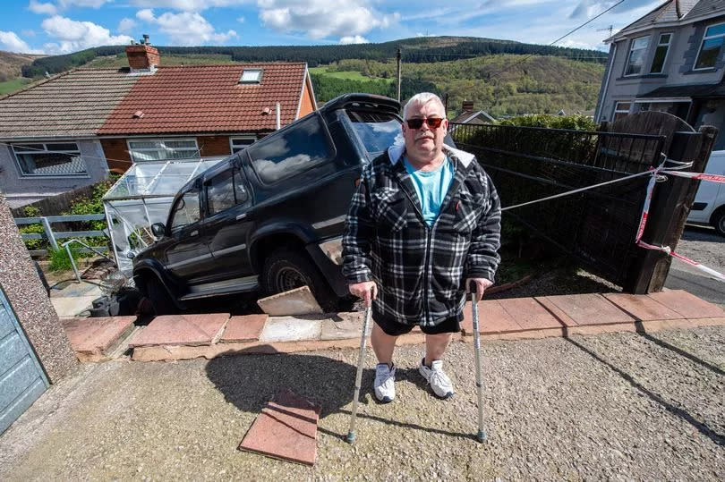 A white man in his 60s, dressed in shorts and wearing sunglasses, holding crutches. He has standing in front of a 4x4 vehicle which is perched at the top end of a garden