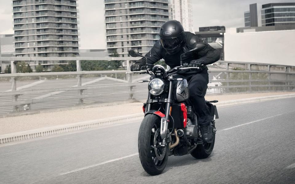 A triumph motorcycle with red detailing is ridden across a road next to high-rise buildings.