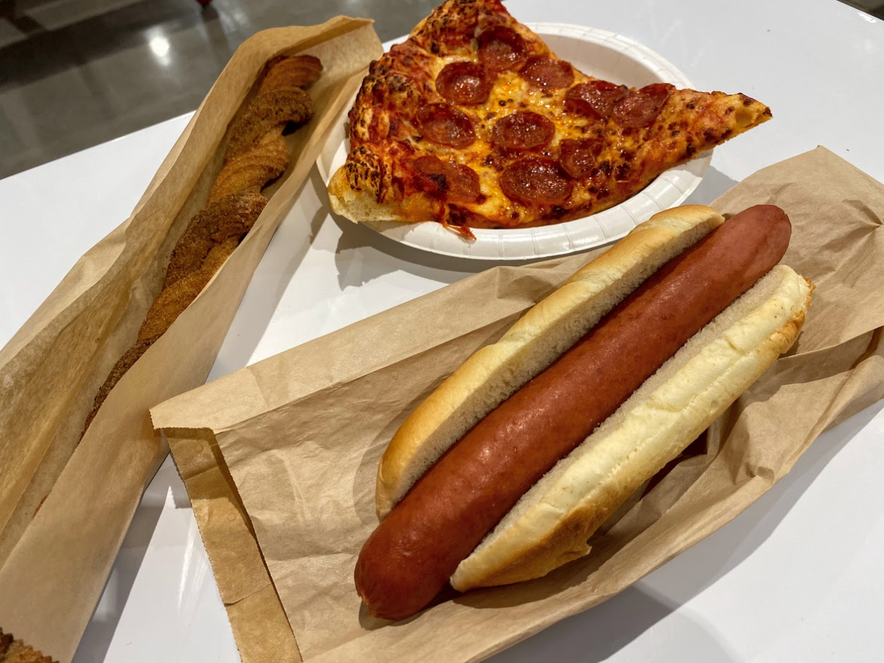 A churro wrapped in brown paper, a slice of pepperoni pizza on a white paper plate, and a hot dog on a brown paper bag, at the Costco food court, on a white table, grey concrete floor in the top left background