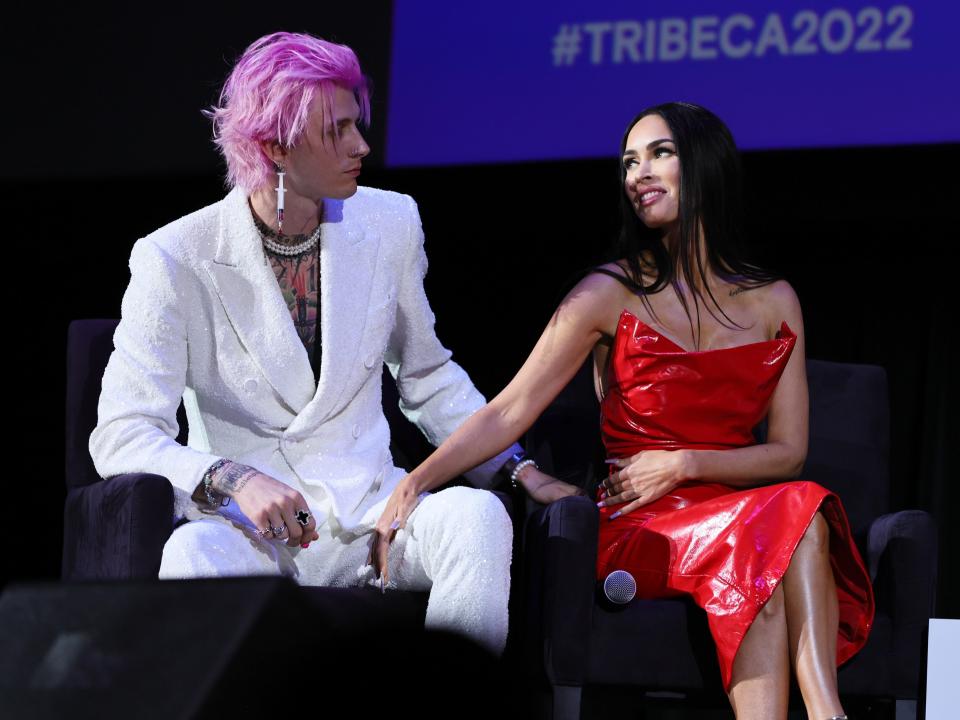 machine gun kelly and megan fox sitting next to each other on stage at the tribeca film festival, looking at each other fondly with fox's hand on kelly's thigh