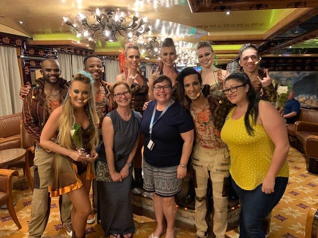 A group of 11 people in two rows posing for a picture in a dining room on a cruise ship. Some are in costumes and makeup with microphones, while others are wearing staff lanyards.