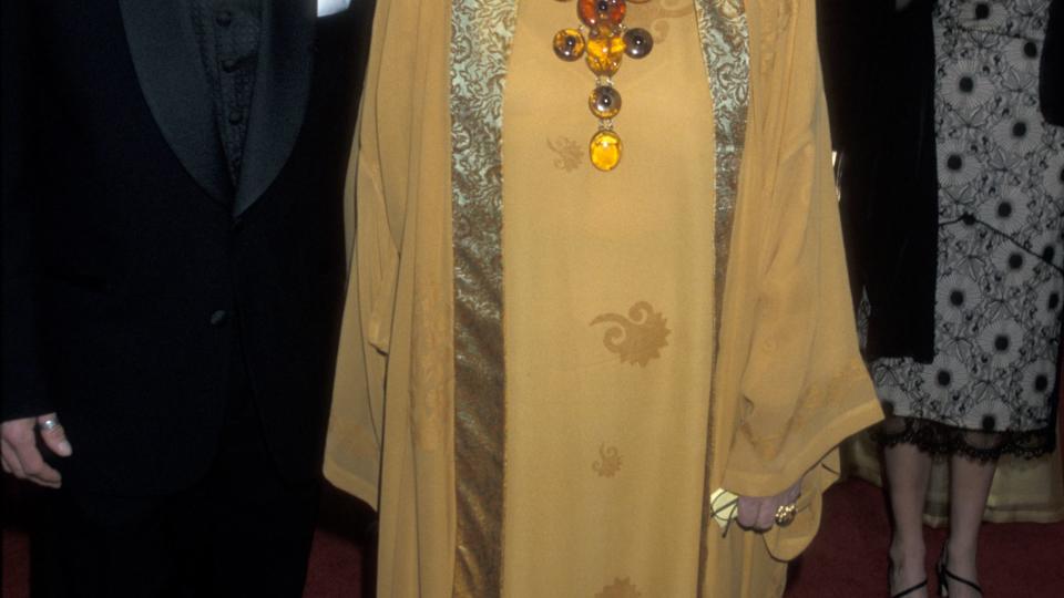 jefferson burstyn and ellen burstyn stand next to each other in front of a group of people, he wears a black tuxedo, she wears a yellow dress and caftan with a gold trim and a large yellow necklace