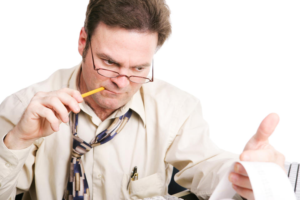 A man with a loosened tie chews on a pencil while closely analyzing figures from his printing calculator.