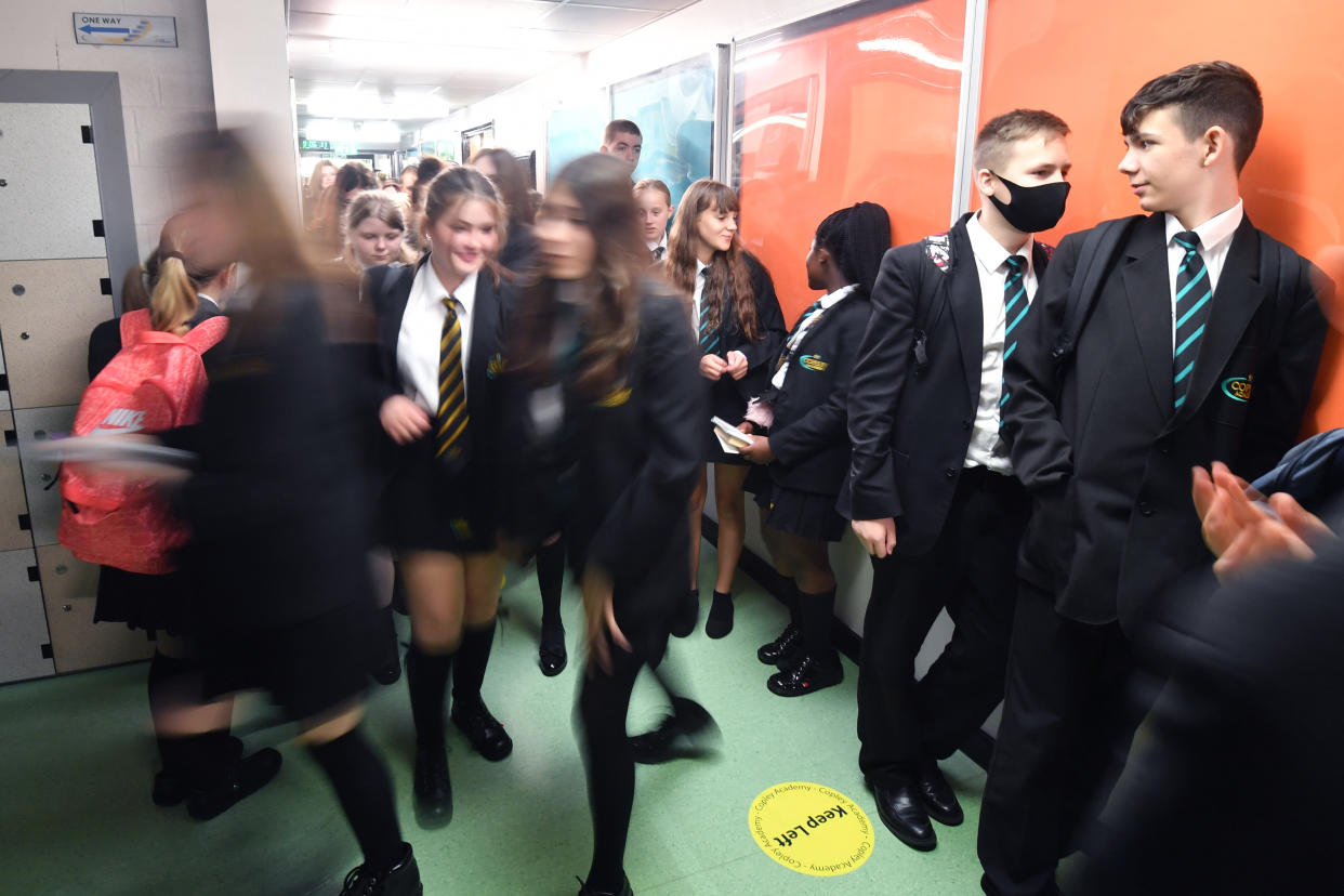 Social distancing signs reading Keep Left are displayed on the floor as students, one of which is wearing a face mask, walk along a school hallway.