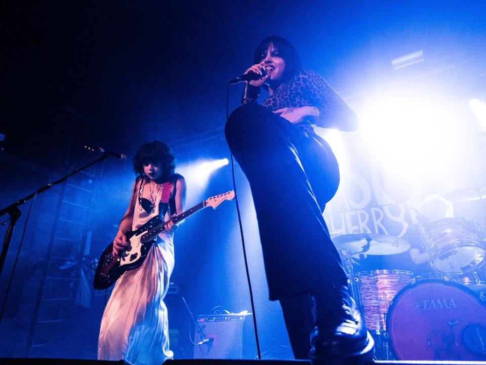 chloe chaidez and gabriette bechtel performing onstage with nasty cherry. the photograph is taken from a low angle, showing chaidez playing the guitar in a white slip dress and bechtel holding a microphone and singing in a cheetah print shirt