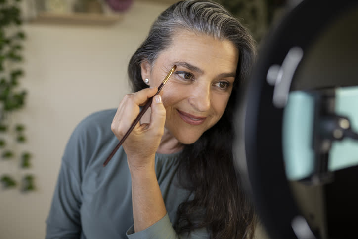 Woman applying makeup using a brush, looking at a ring light mirror