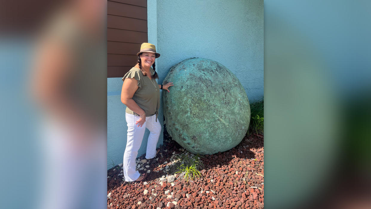  We see a woman standing outside a building next to a large circular green drum-shaped object.  