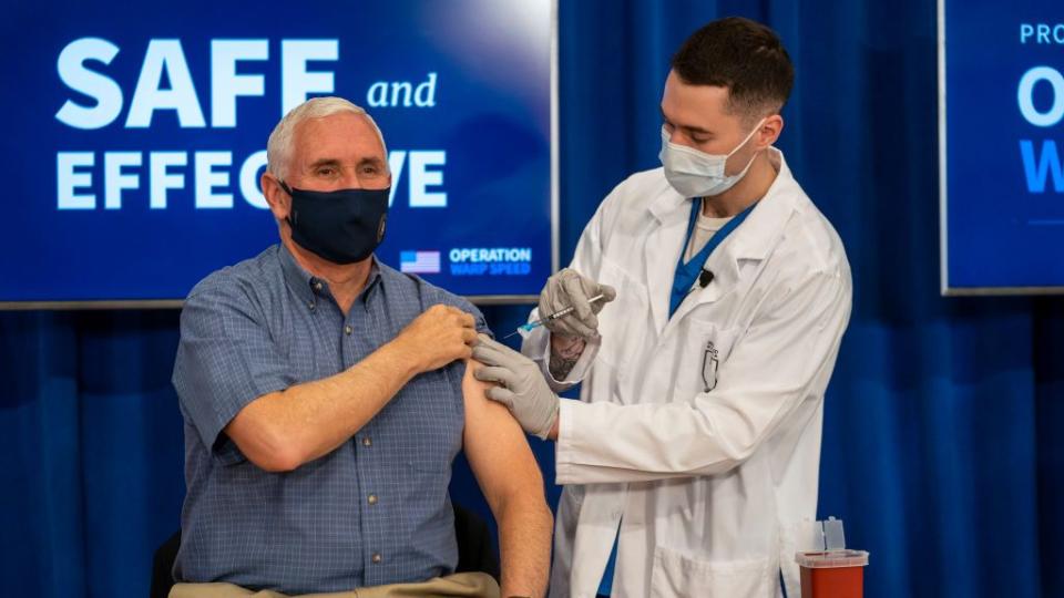 vice president pence is sitting down and hold his left sleeve shirt as a health care provided sticks a vaccination needle into his arm, both men are wearing face masks