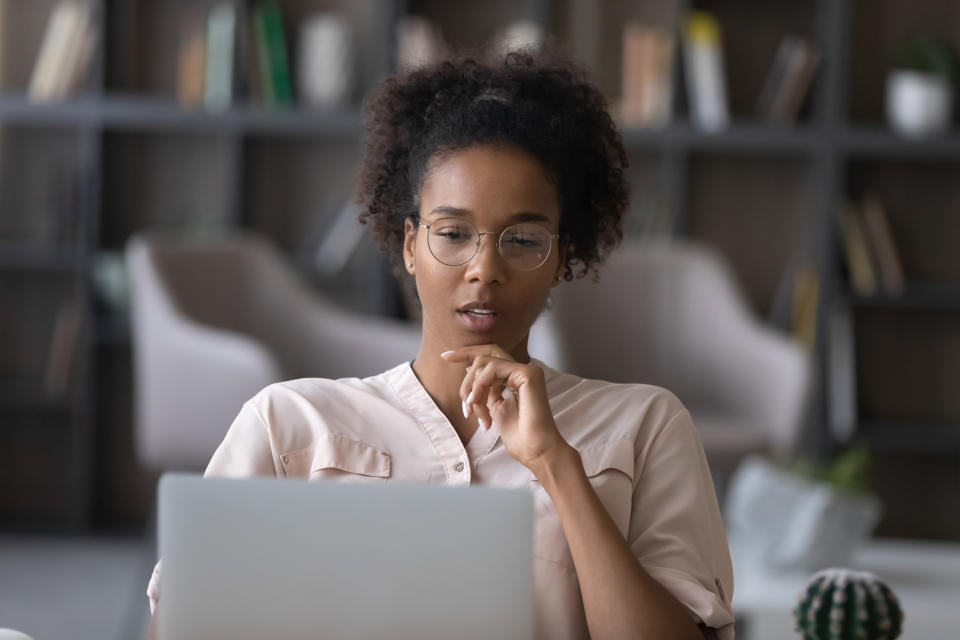 Thoughtful young African American woman in glasses look at laptop screen work online at home office.