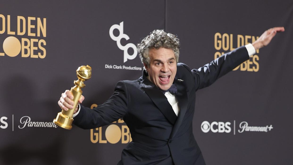 mark ruffalo smiles and jumps on a red carpet while holding a golden globe trophy in one hand, he wears a black tuxedo and appears in front of a black background with logos on it