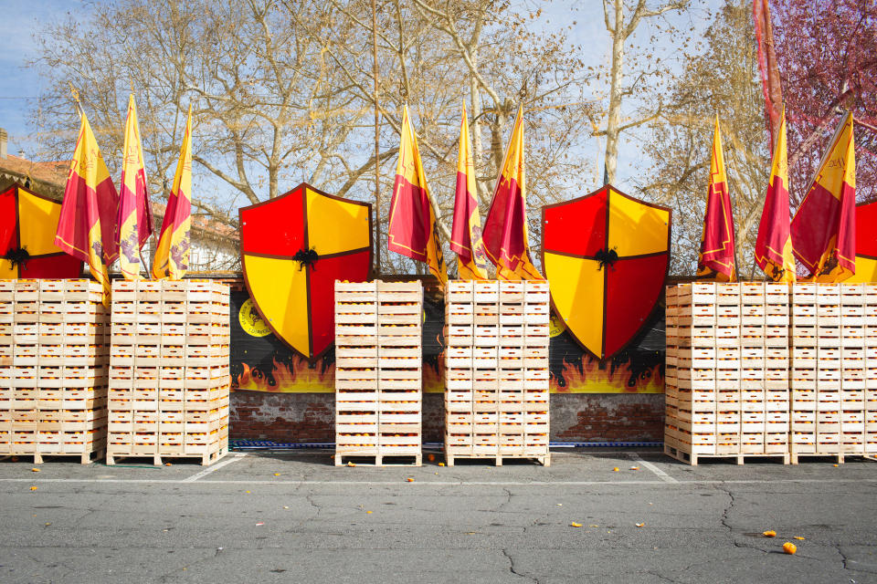 Six tall, vertical stacks of wooden crates of oranges sit in a parking lot in front of a coat of arms and flags depicting the team's colors