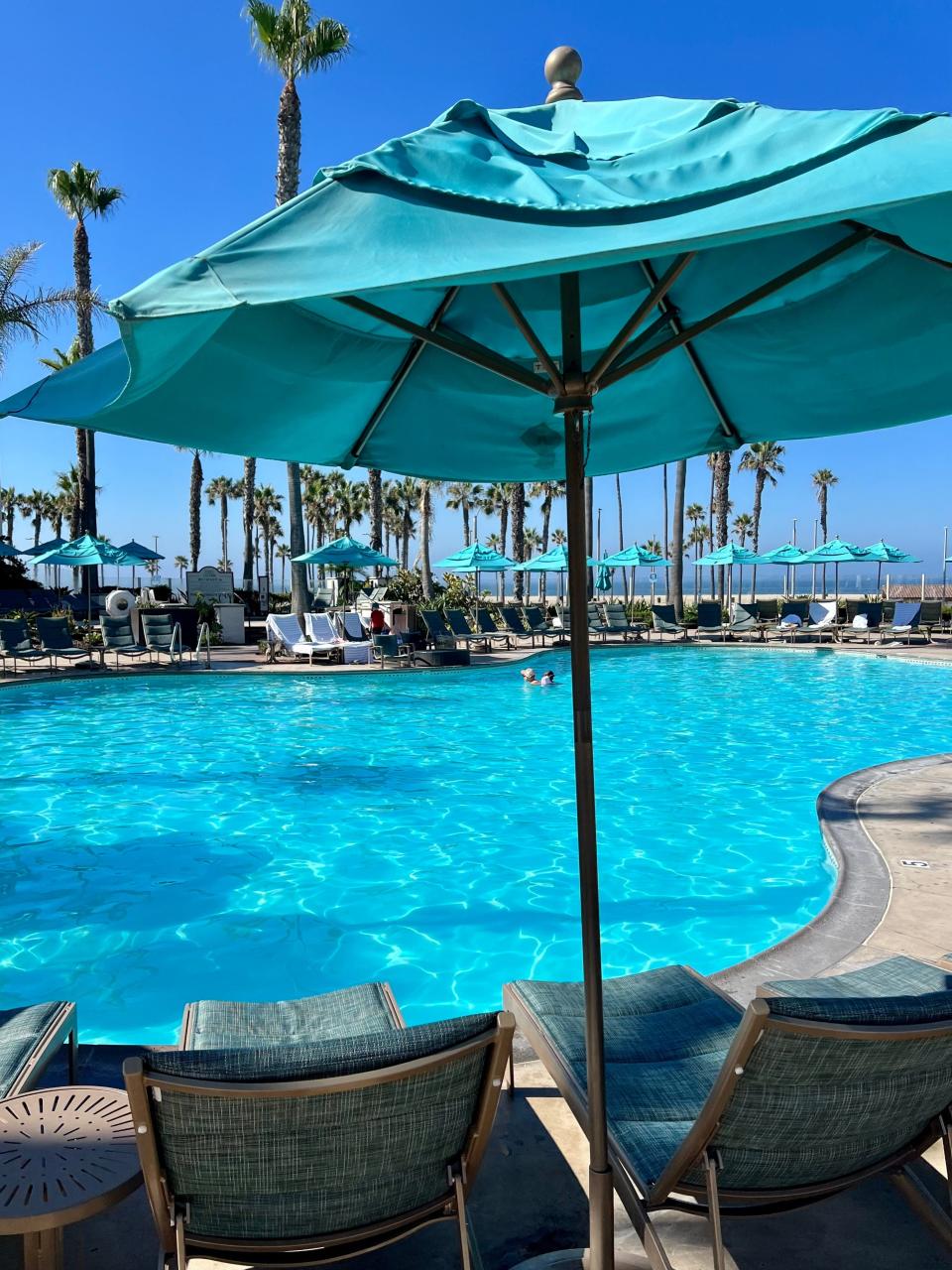 Empty pool chairs under a blue umbrella facing a blue lagoon-shaped pool.