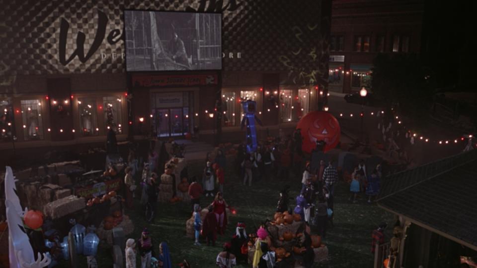 A wide shot of a town square decorated for Halloween with many people in costumes, as an old black and white movie plays on a screen in the background.