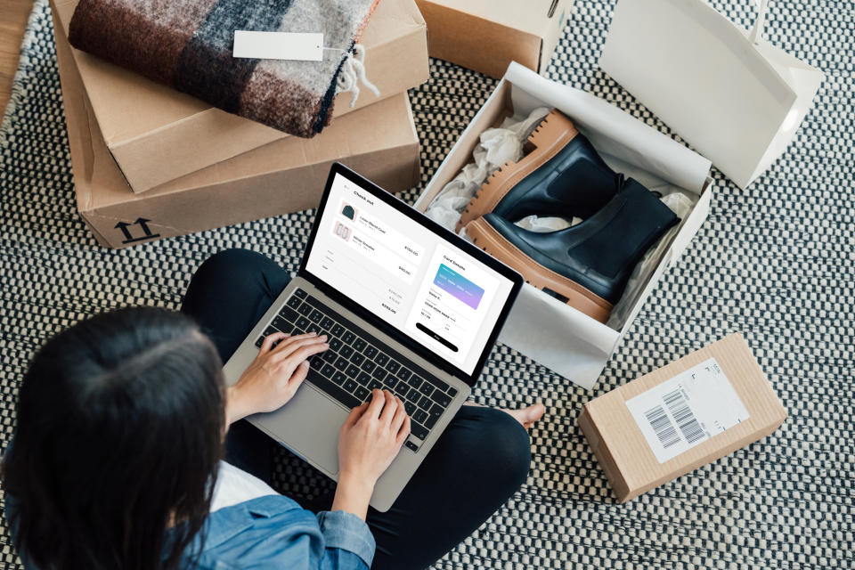 A person sits on a rug with a laptop, surrounded by open shopping boxes including shoes and a checkered blanket, suggesting online shopping