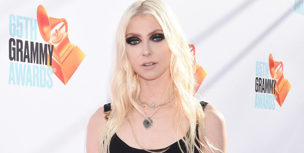 taylor momsen, a young woman stands looking at the camera, she has long blonde hair worn down in loose waves, heavy black eye makeup and wears a short black dress