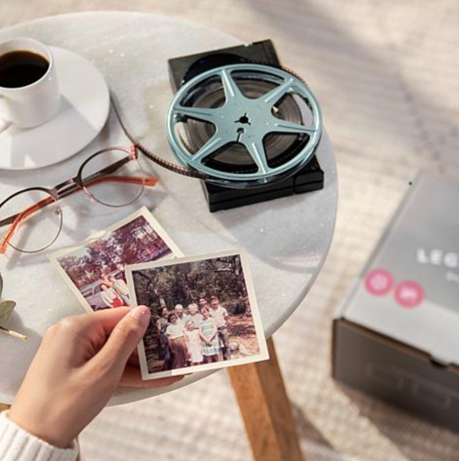 Woman holding old photos above a table with film reels and a Legacybox on the floor. 