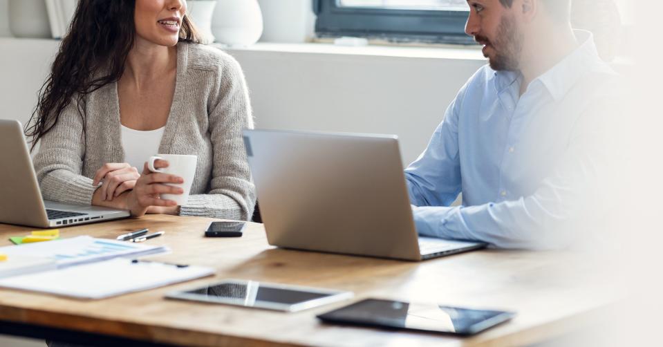 Two people sit at a table using laptops and talking with each other in a modern office setting