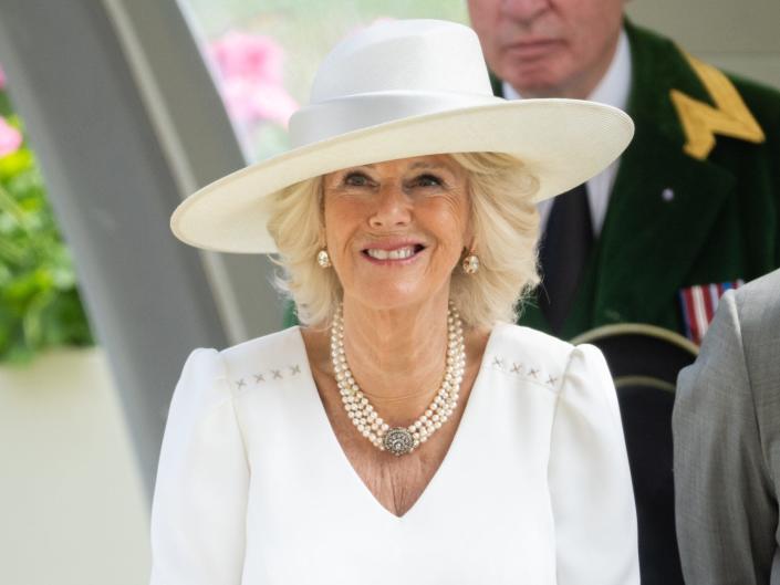 Camilla Parker Bowles smiles in a white dress and white hat.