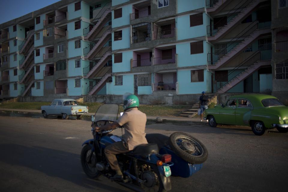 In this April 12, 2014 photo, a man drives his motorcycle with a sidecar next to buildings of the Alamar neighborhood in Havana, Cuba. Around Havana, Cubans can be seen taking advantage of the materials now available as they add second stories to their homes, enclose balconies to turn them into an extra room or throw on a fresh coat of paint. (AP Photo/Ramon Espinosa)
