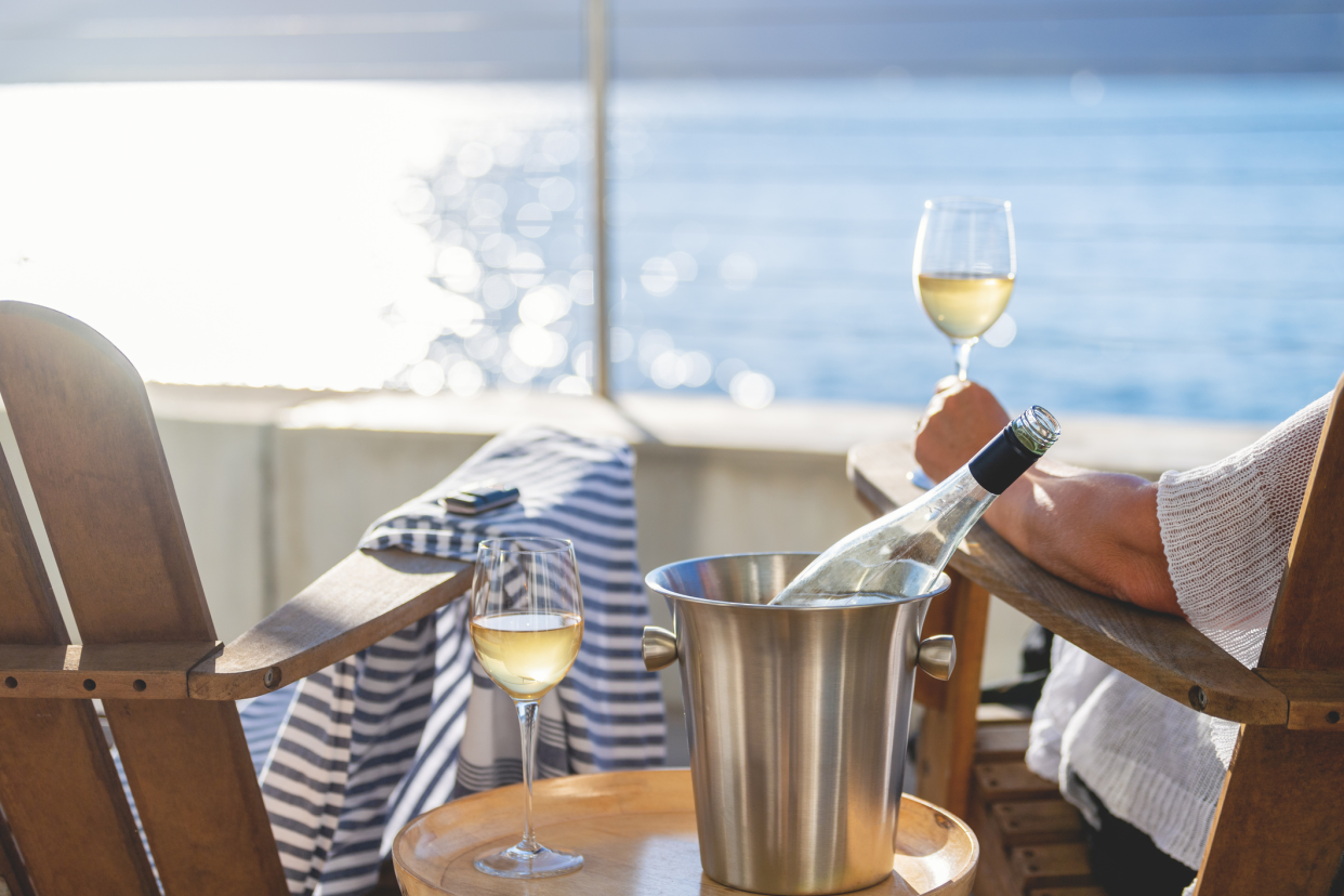 Two deck chairs on the balcony of cruise, woman sitting in one of the chair enjoying a glass of white wine, ice bucket with wine bottle and another glass, ocean and sunlight on waves blurred in the background