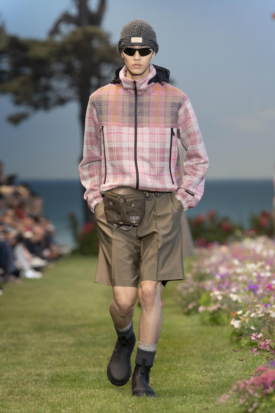 Longer trouser shorts paired with a zip-up jacket, fanny pack, beanie and rubberized boots at Dior summer ’23 men’s. - Credit: YANNIS VLAMOS