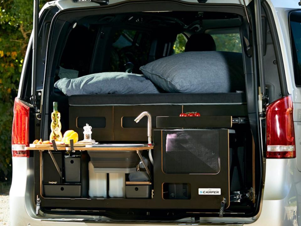 The trunk of the EQV open, revealing a mini kitchen. The van is parked next to an outdoor dining set.