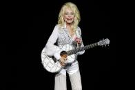 <p>Dolly Parton dazzles the crown with an all-white ensemble (and matching guitar!) in Perth, Australia. Her uniform is often a simple blouse with a beaded vest and pants for comfortable glamour.</p>