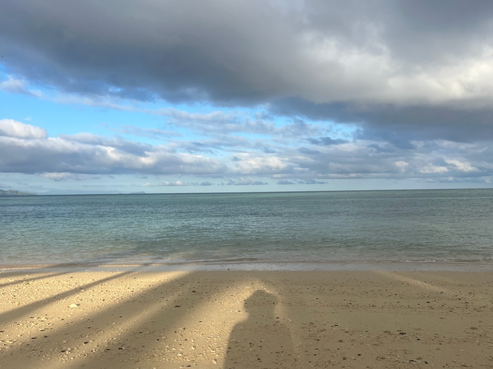 Beachfront covered in tan sand, blue waters, and clouds among blue sky. A shadow of the writer is in the foreground. Some clouds are dark