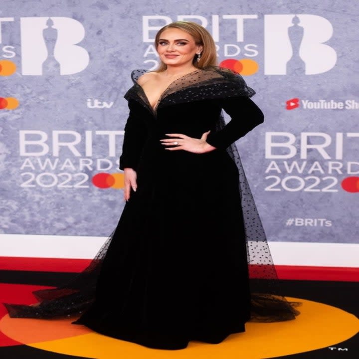 Adele poses on the red carpet with a hand on her hip while rocking an elegant off-the-shoulder, long-sleeved gown