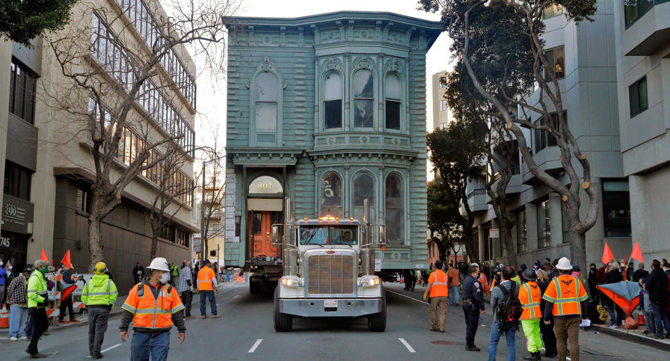 Dozens of tradesmen are seen flanking the house being moved on a truck.