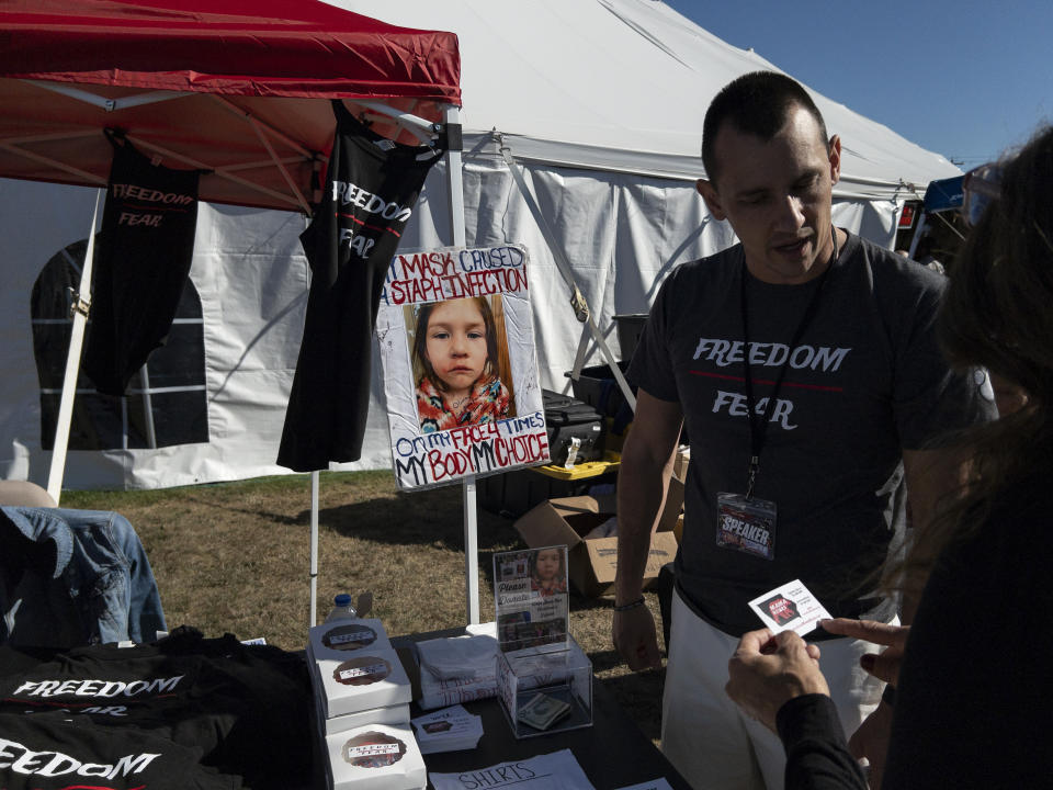 Jonathan Reicks talks at Iowa Mama Bears’ “Freedom Over Fear” merchandise table during the ReAwaken America Tour at Cornerstone Church, in Batavia, N.Y., Friday, Aug. 12, 2022. A poster bearing a photo with Reicks’ kindergarten-aged daughter is displayed at their booth. (AP Photo/Carolyn Kaster)