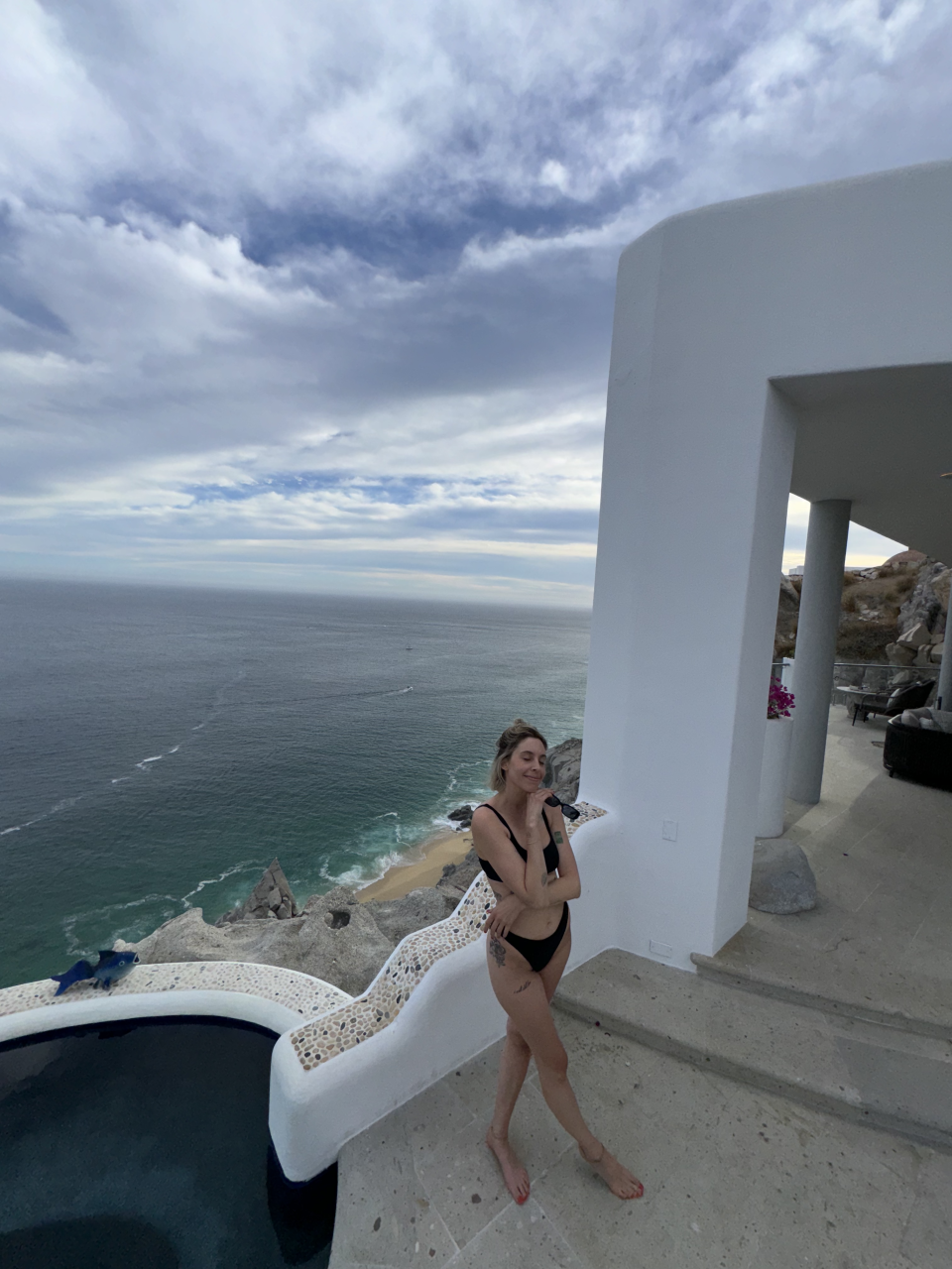 A woman in a black bikini poses near an infinity pool with an ocean view from a coastal cliffside villa