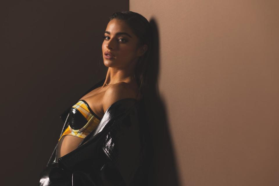 Noa Kirel wears a bikini top and leather jacket as she leans against a wall.