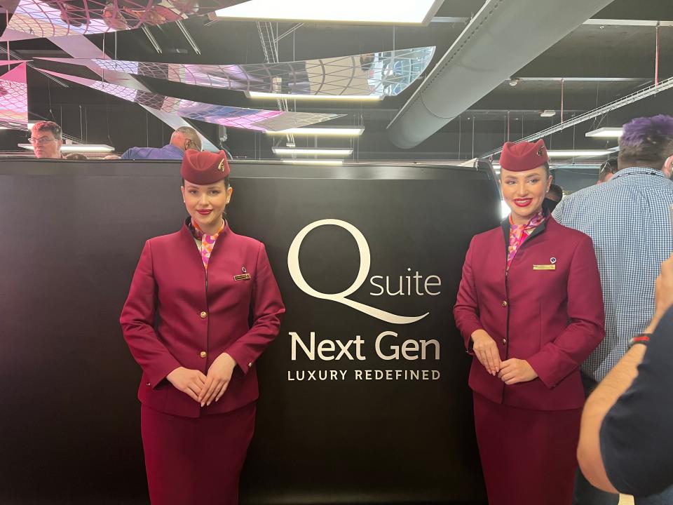 Two Qatar Airways flight attendants stand in front of the QSuite Next Gen display at the Farnborough Airshow 2024.