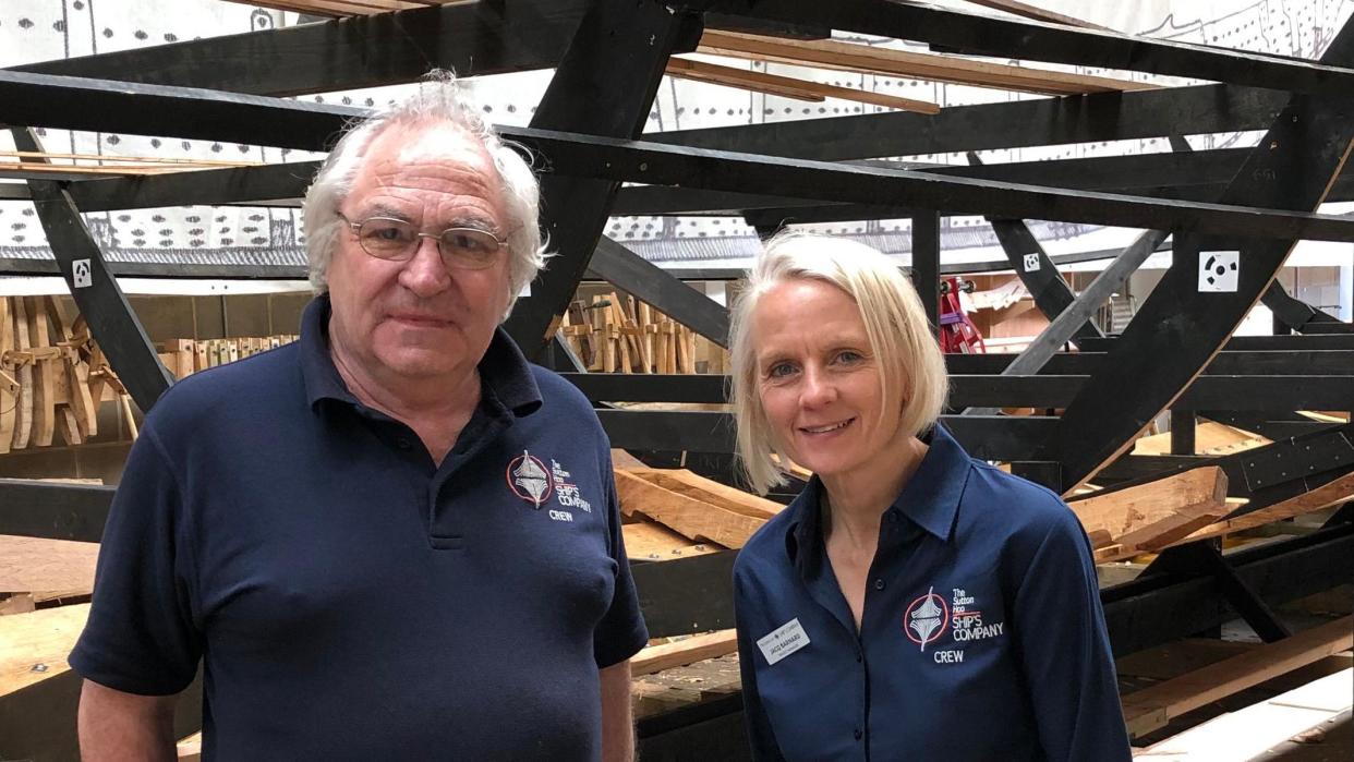 Tim Kirk and Jacq Barnard both in dark blue Ships Company tops standing in front of frame with half built replica Sutton Hooship