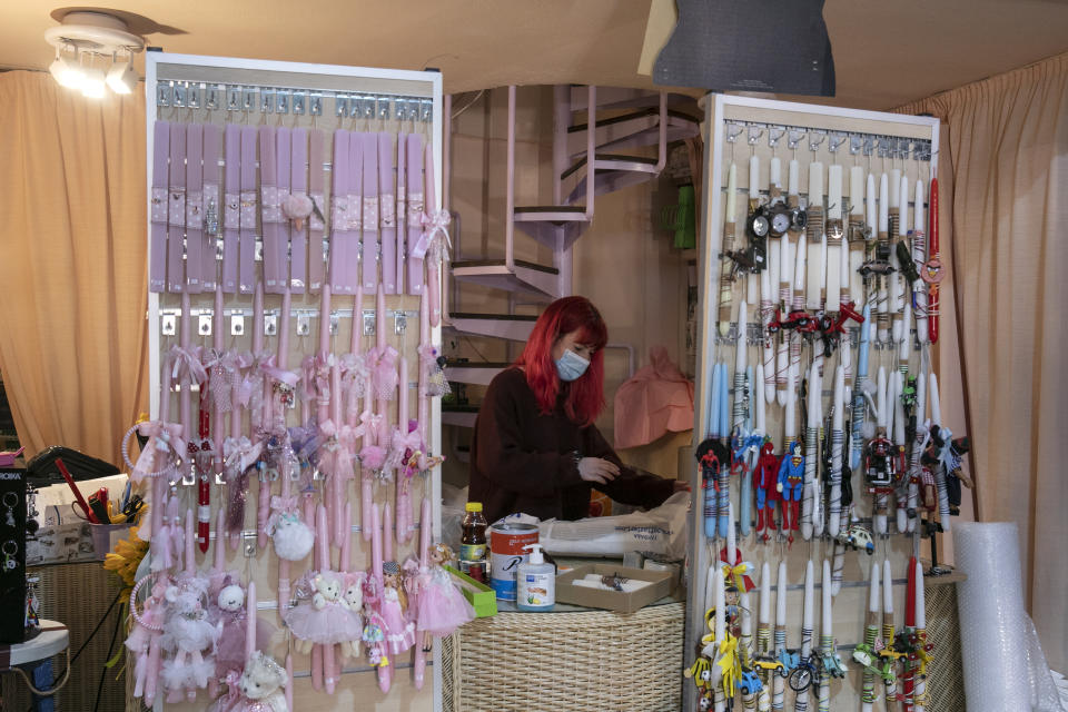 An employee, wearing a face mask to curb the spread of the coronavirus, works behind decorated candles for the Orthodox Easter, inside a small gift shop, at Glyfada suburb west of Athens, Saturday, April 3, 2021. Greece has relaxed some coronavirus restrictions despite surging COVID-19 cases that are straining hospitals to their limits, with retail stores to reopen and people allowed to drive outside their home municipalities for exercise on weekends. (AP Photo/Yorgos Karahalis)