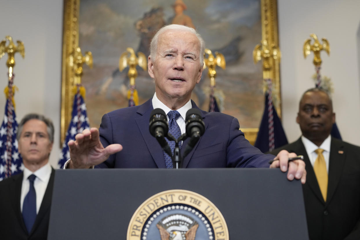 President Biden speaks from a podium as Secretary of State Antony Blinken and Defense Secretary Lloyd Austin stand behind him.. 