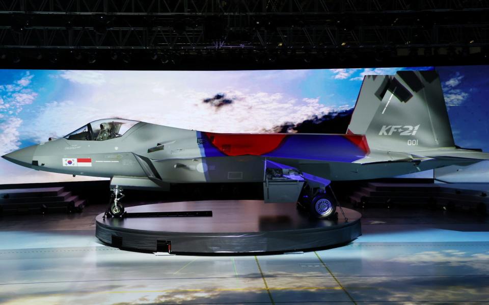 A South Korea KF-21 fighter jet on a circular stand in front of a screen displaying a cloudy sky at a ceremony.