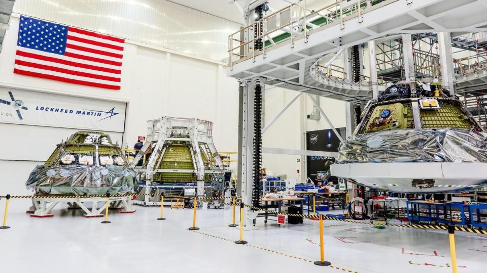 three cone-shaped spacecraft beside each other in a big white hanger. the american flag and the logo for lockheed martin are on the wall