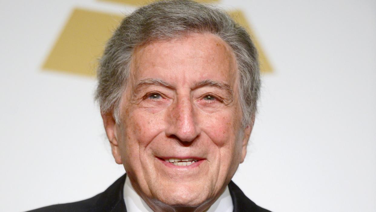 tony bennett smiles for a photo, he wears a black suit and tie with a red pocket square, part of a grammy award is visible in the foreground